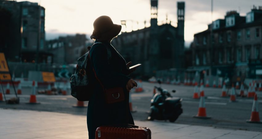 woman wearing coat standing on road with travel luggage during daytime